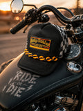 Hempin Mayhem black trucker hat with checkered mesh, yellow and black chain detail, resting on a motorcycle tank.