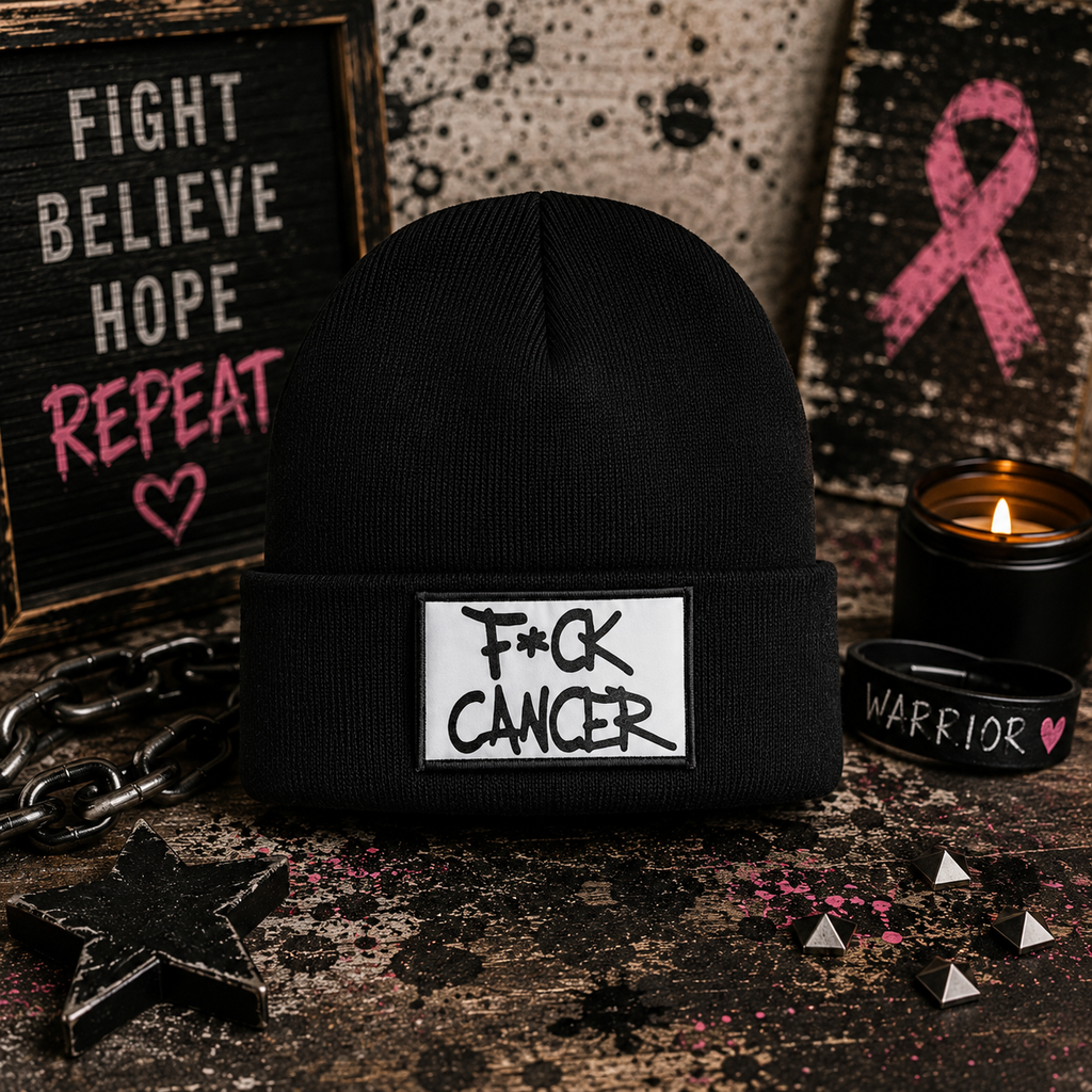 A black knit beanie with a white rectangular patch reading 'F*CK CANCER' in black lettering, styled on a dark tabletop with a candle, bracelet, chain, and pink ribbon wall art in the background.