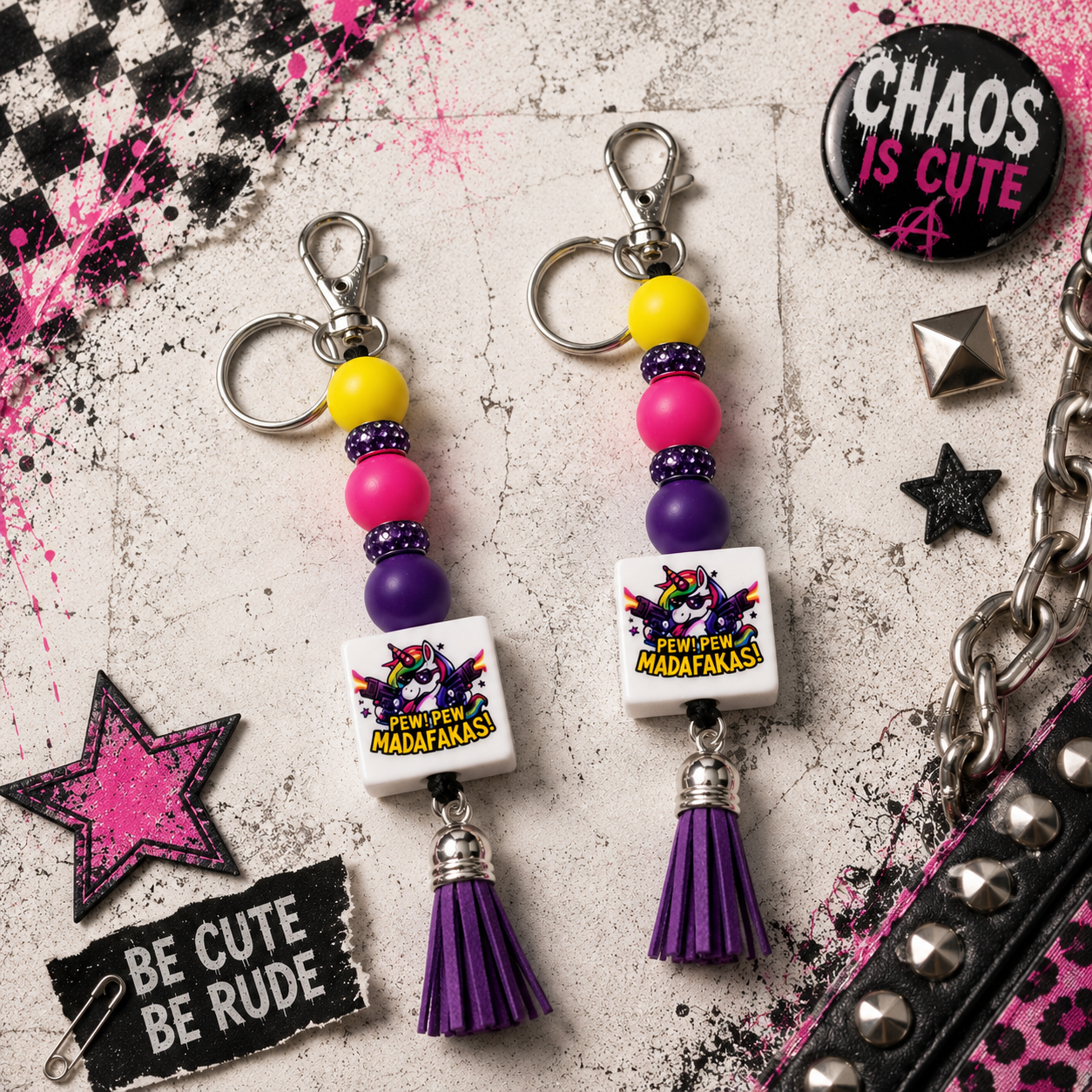 Two matching keychain beaded charms with silver keyrings, colorful beads, white square pendants printed with Madafanaka graphics, and purple tassel charms, arranged on a distressed light gray background with pink and black decorative accessories.