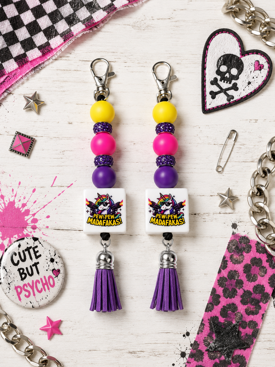 A pair of beaded dangle earrings with purple tassels, displayed on a white wood background surrounded by keychain accessories like a skull patch, safety pin, and charm.