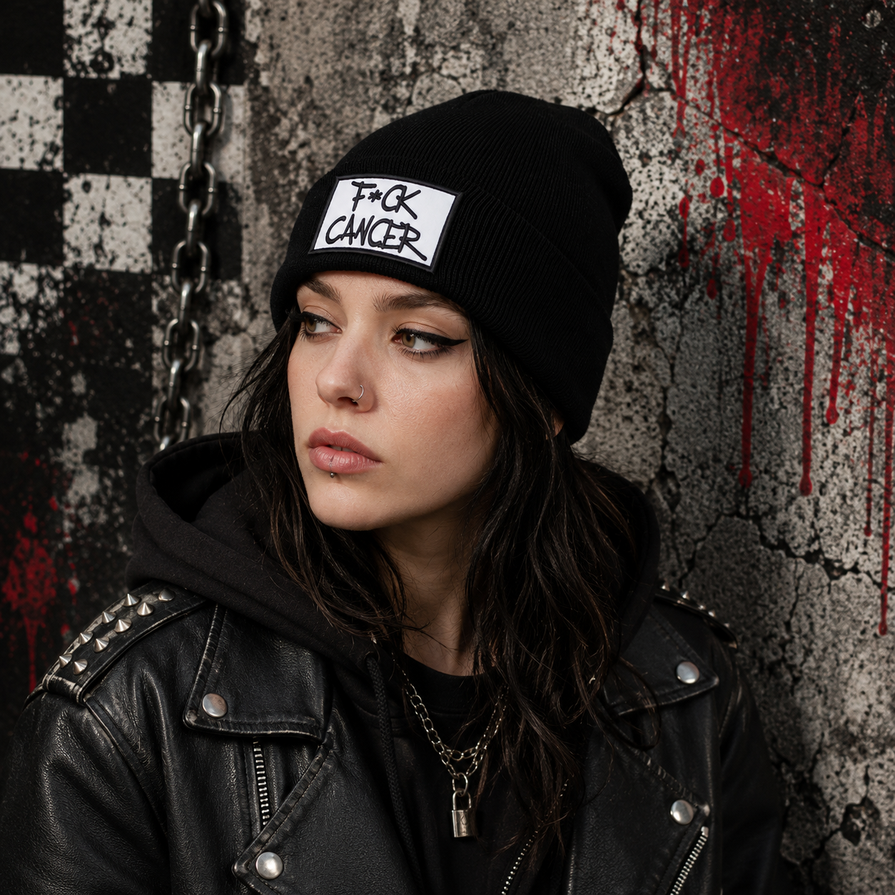 A woman wearing a black beanie with a white patch that reads 'F*CK CANCER' and a black leather jacket, posed against a distressed concrete wall with a black-and-white checkered pattern and red paint drips.