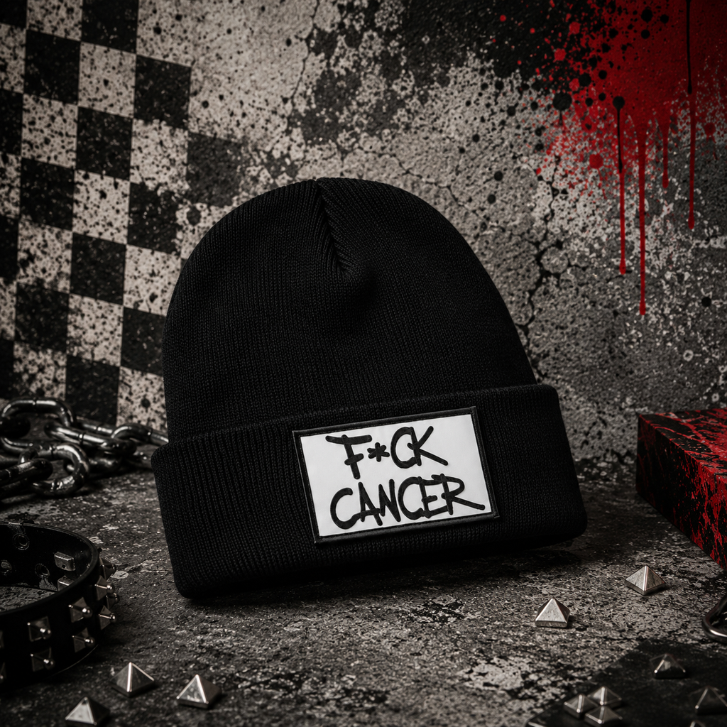 A black knit beanie with a white rectangular patch on the cuff that reads 'F*CK CANCER' in black handwritten-style text. The beanie is photographed in a styled studio setup with a distressed black-and-white backdrop, red paint drips, chain, studs, and other dark props.