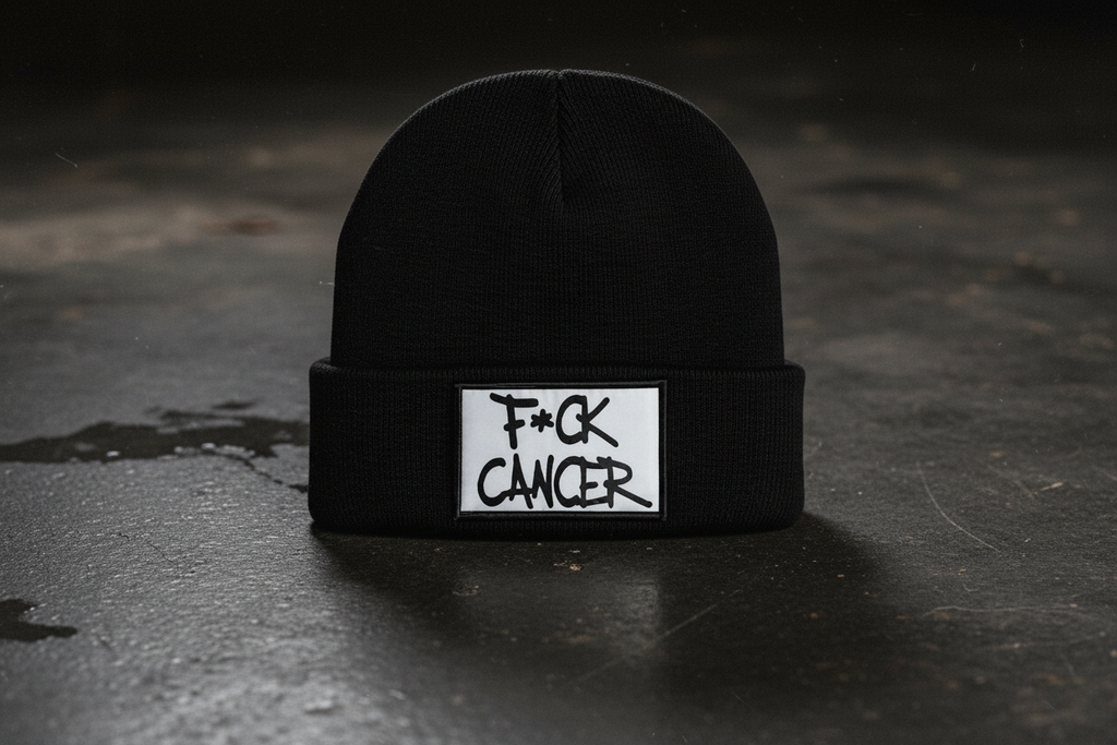 A black knit beanie hat with a white rectangular patch on the folded cuff that reads 'F*CK CANCER' in black handwritten-style text, photographed on a dark textured surface.