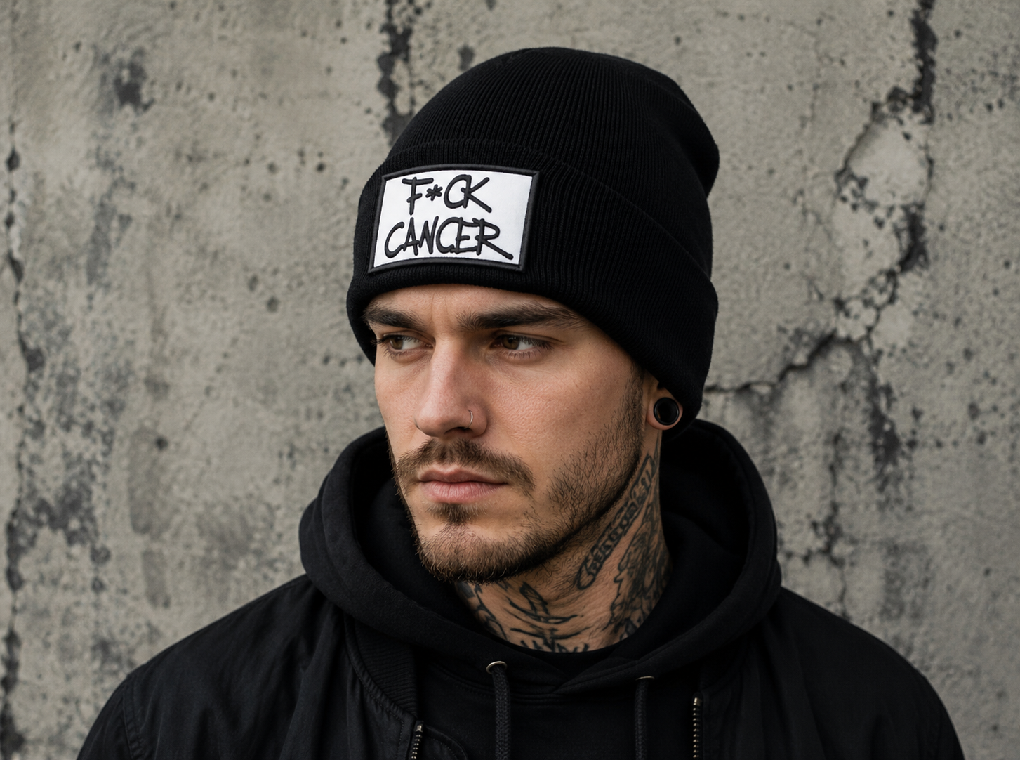 A man wearing a black knit beanie with a white rectangular patch that reads 'F*CK CANCER' in black text, posed against a textured gray concrete wall.